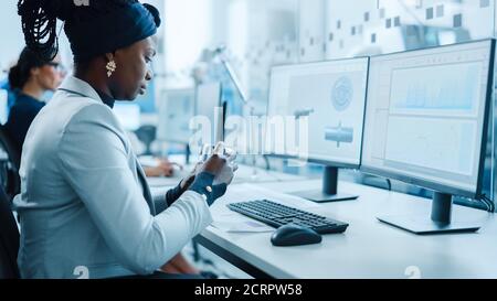 Beautiful Female Engineer Working on Personal Computer in the High-Tech Industrial Factory, She Uses Smartphone for CAD Software Designing 3D Stock Photo