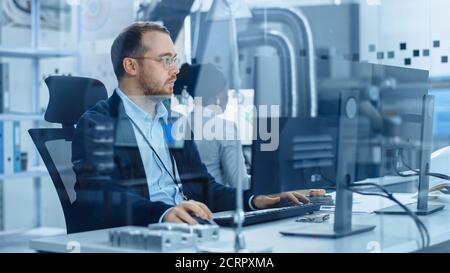 Factory Office: Industrial Engineer working on Personal Computer, On his Desk Lying Various Blueprints, Electronic Mechanisms, Heavy Industry Stock Photo