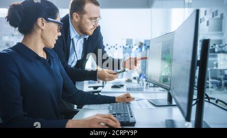 Modern Electronics Factory: Male Supervisor Talks to a Female Electrical Engineer who Works on Computer with CAD Software. Developing PCB, Microchips Stock Photo