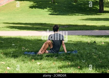 Young athletic man in sportswear doing yoga in the park. Practice half spinal twist asana outdoors. People exercising stretching on green grass with Stock Photo