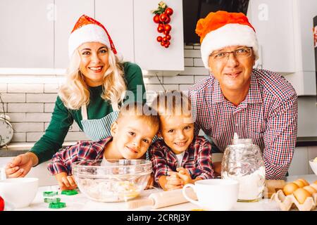 Family of two twins boys and age parents preparing cookies for Chtistmas in light kitchen wearing Santa hats and looking at camera Stock Photo