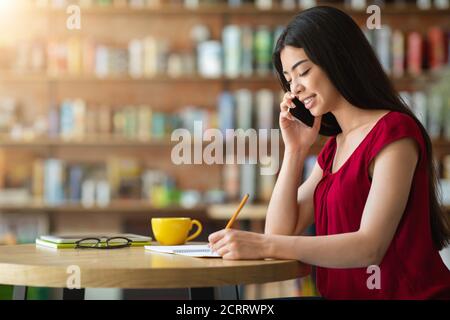 Smiling Asian Woman Talking On Cellphone And Taking Notes In Cafe Stock Photo