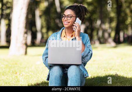 Smiling Black Student Woman Speaking On Cellphone Using Laptop Outdoors Stock Photo