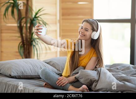 Happy young lady in headphones taking selfie with mobile device on bed at home Stock Photo