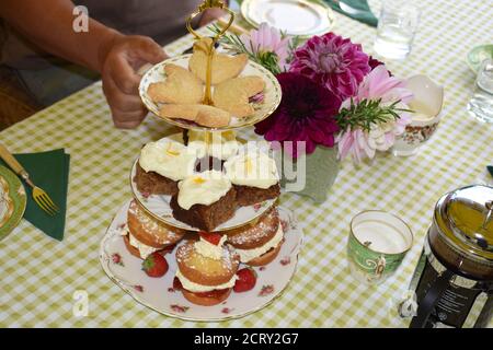 Homemade afternoon tea related ritual served with hot drinks. Cakes stand featuring shortbread courgette and mini Victoria sponge strawberry jam cake. Stock Photo