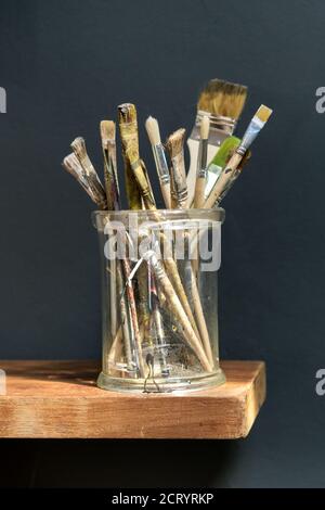 Glass jar filled with assorted used artists paintbrushes with stained tips to the bristles on a wooden shelf Stock Photo