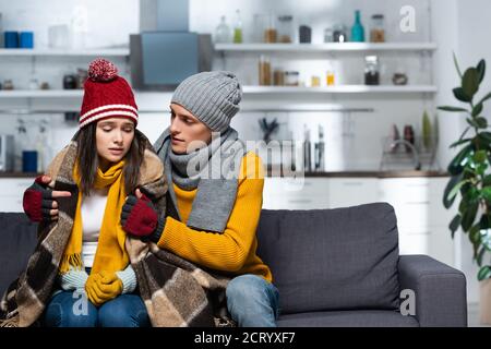 young man in warm hat and gloves wrapping cold girlfriend with plaid blanket while sitting in kitchen Stock Photo
