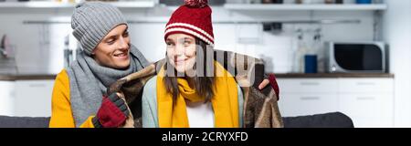 horizontal image of man in knitted hat wrapping joyful girlfriend with warm plaid blanket while sitting in cold kitchen Stock Photo