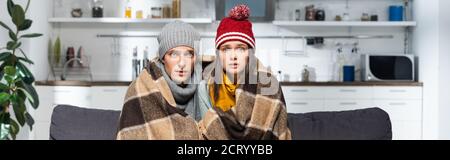 panoramic crop of freezing couple, wearing warm hats, looking at camera while sitting in cold kitchen under plaid blanket Stock Photo