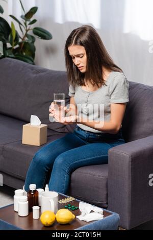 diseased woman holding glass of water and pills while sitting near bedside table with medicines and fresh lemons Stock Photo