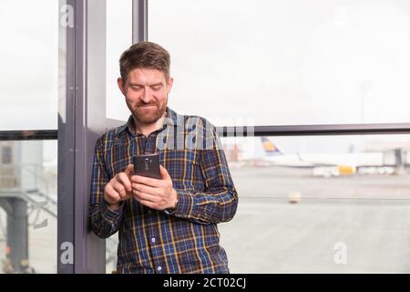 Happy man browsing smartphone while waiting for flight near window in airport Stock Photo