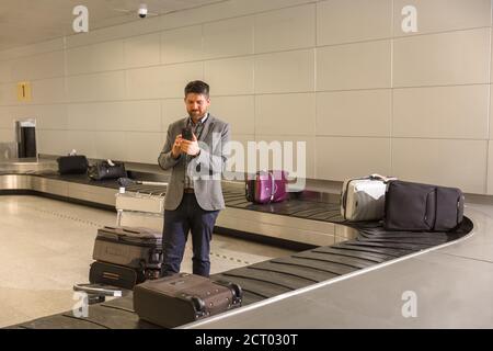 Male traveler standing near luggage carousel and browsing smartphone in airport Stock Photo
