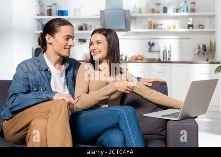 man sitting near excited girlfriend pointing with finger at laptop on couch in kitchen Stock Photo