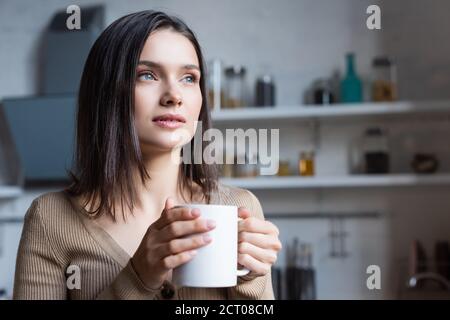 dreamy brunette woman looking away while holding cup of tea at home Stock Photo