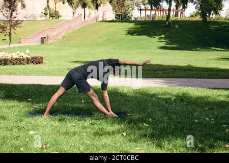 Young athletic man in sportswear doing yoga in the park. Practice triangle asana pose outdoors. People exercising on green grass with yoga mat Stock Photo