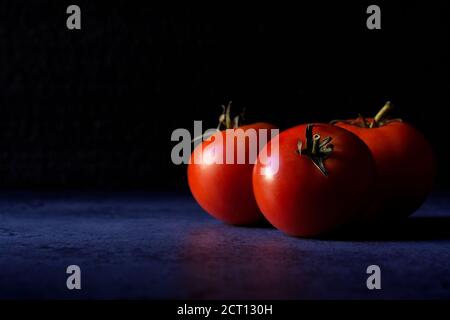Three mature tomato fruits are lying on a dark blue background. There is a lot of copy space beside them. Selective focus. Stock Photo