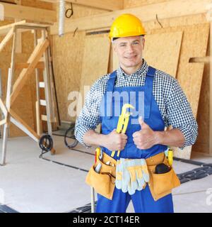 Happy male construction worker in blue uniform coveralls and yellow hardhat holding wrench showing thumb up and smiling Stock Photo