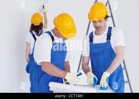 Building, renovation, repair, teamwork and people concept - close up of builders discussing blueprint indoors woman painting wall on backround Stock Photo