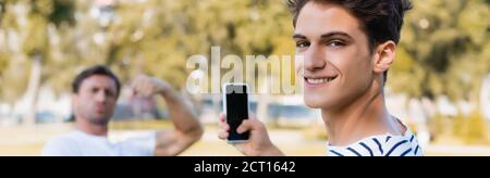 horizontal concept of joyful teenager boy taking photo of father while holding smartphone in park Stock Photo