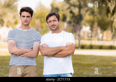 father and teenager son standing with crossed arms in park Stock Photo