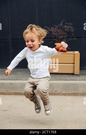 Playful little boy with blond hair jumping on the street with croissant in hand