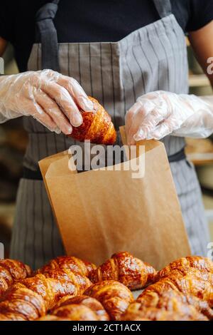 Careful baker placing freshly baked crispy golden croissants in a paper bag