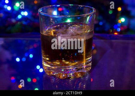 Glass with whiskey and ice cubes on blue background. The Christmas tree is lit in the background. Glass is mirrored on a glass table. Stock Photo