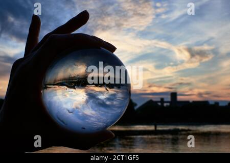 Silhouette of a female hand holding a lensball in front of the beautiful sunset. Dramatic cloudy sky above the water on a summer evening. Stock Photo