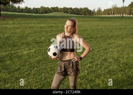 Soccer girl. Beautiful sporty young woman holding soccer ball and smiling at camera while standing outdoors in a green field Stock Photo