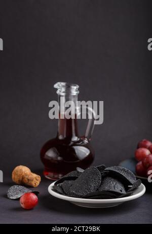 Black potato chips with charcoal, balsamic vinegar in glass, red grapes on a blue ceramic plate on a black background. side view, close up, selective Stock Photo