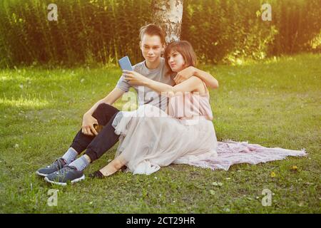 Young attractive man and woman 23 years old is taking selfie by smartphone while sitting on the grass in the public park. Stock Photo