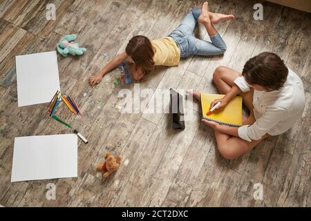 Brother and sister lying on the wooden floor at home and spending time together. Little girl playing with puzzles, boy using digital tablet and