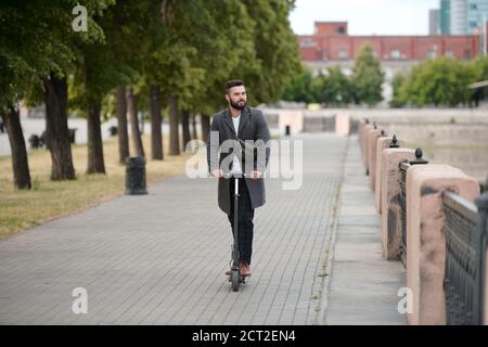 Contemporary young bearded businessman in coat riding on electric scooter Stock Photo