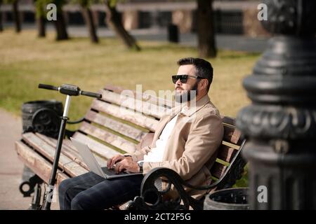 Young pensive businessman in smart casualwear and sunglasses using laptop Stock Photo