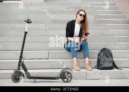 Contemporary female student with glass of coffee sitting on staircase outdoors Stock Photo