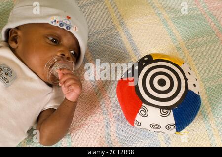 6 week old newborn baby boy one month premature alert looking at toy with high contrast black and white pattern, wearing cap and sucking on pacifier Stock Photo