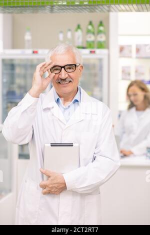 Successful senior drugstore worker in whitecoat and eyeglasses using tablet Stock Photo