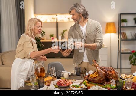Mature man in casualwear showing bottle of wine to his wife over served table Stock Photo