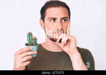 Young handsome man holding small cactus pot serious face thinking about question with hand on chin, thoughtful about confusing idea Stock Photo