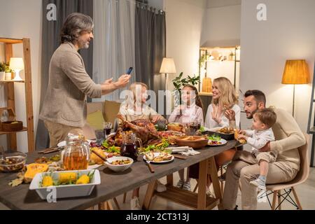 Cheerful mature man with smartphone taking photo of his big family by dinner Stock Photo