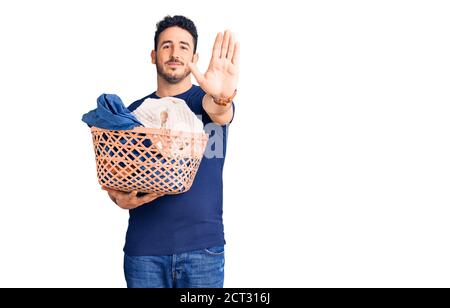 Young hispanic man holding laundry basket with open hand doing stop sign with serious and confident expression, defense gesture Stock Photo