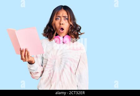 Young beautiful mixed race woman reading a book wearing headphones scared and amazed with open mouth for surprise, disbelief face Stock Photo