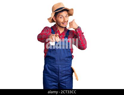 Handsome latin american young man weaing handyman uniform smiling doing talking on the telephone gesture and pointing to you. call me. Stock Photo