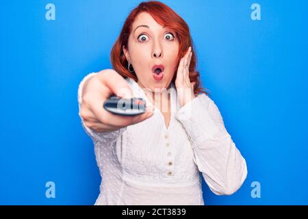 Young beautiful redhead woman changing television channel using tv remote control scared and amazed with open mouth for surprise, disbelief face Stock Photo