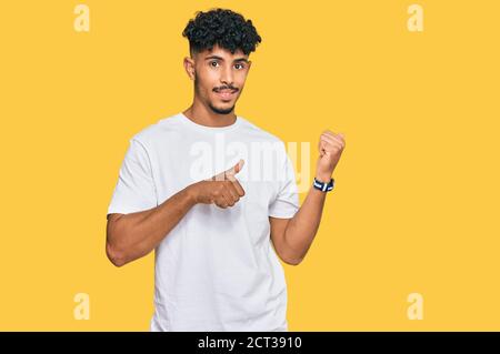 Young arab man wearing casual white t shirt pointing to the back behind with hand and thumbs up, smiling confident Stock Photo