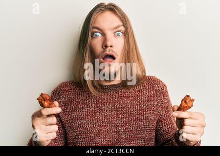 Handsome caucasian man with long hair eating chicken wings afraid and shocked with surprise and amazed expression, fear and excited face. Stock Photo