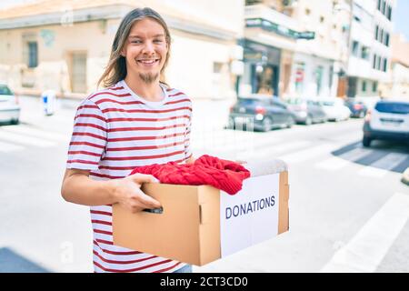 Young caucasian man with blond long hair and beard holding cardboard box with clothes from donations Stock Photo