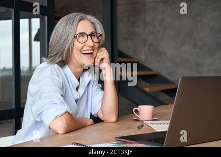 Happy mature middle aged woman laughing sitting at workplace with laptop. Stock Photo