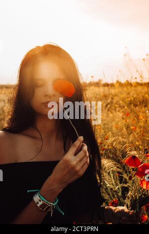 Summer portrait of beautiful tanned, brunette Ukrainian woman with long dark hair closing one eye with blooming red poppy and bouquet in hand in off s Stock Photo