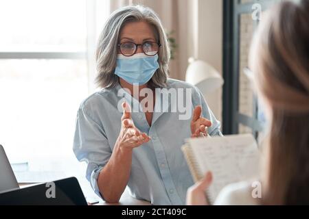 Old female mentor wearing face mask training young interns at office meeting. Stock Photo